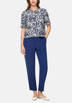 Frau trägt ein kurzärmeliges, marineblau-weiß gemustertes Oberteil, königsblaue Hose und weiße spitz zulaufende Ballerinas, steht mit einer Hand in der Tasche.