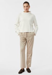 Cremefarbener Rollkragenpullover mit geripptem Saum, kombiniert mit beigen, maßgeschneiderten Hosen mit Kordelzug und gesteppten Akzenten. Braune Slipper.