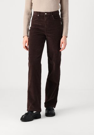 Persona con pantalones de pana marrón oscuro de cintura alta, suéter beige acanalado, zapatos tipo mocasín negros, de pie contra un fondo blanco liso.