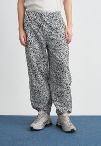 Személy laza, fekete-fehér pettyes nadrágot visel, amely rugalmas mandzsettával rendelkezik, valamint bézs sportcipőt, kék szőnyegen áll.