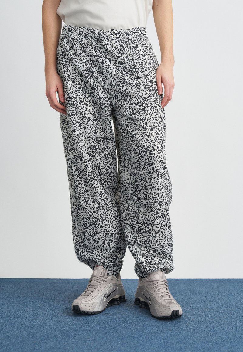 Személy laza, fekete-fehér pettyes nadrágot visel, amely rugalmas mandzsettával rendelkezik, valamint bézs sportcipőt, kék szőnyegen áll.