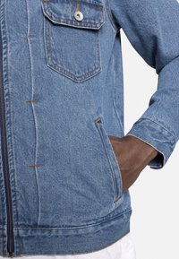 Jeansjack in een lichte blauwe kleur, met zichtbare stiksels, een voorzak en een contrasterende bruine binnenvoering bij de mouw.