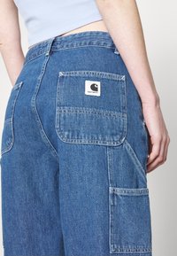Blå denim cargo bukser har en avslappet passform, flere lommer, og et Carhartt-logo merke på baksiden. Stoffet har en teksturert finish.