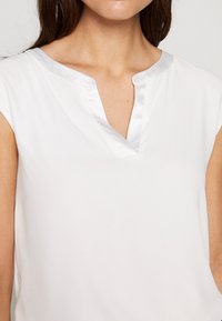 Weiße Bluse mit einem Satinbesatz rund um einen V-Ausschnitt, kurzen Kappenärmeln und glatter Stofftextur. Einfaches und elegantes Design.