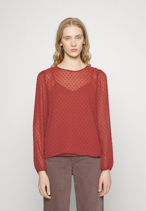 Blusa vermelha transparente com pontos texturizados, gola redonda e mangas longas franzidas nos punhos, combinada com calças de denim castanho.