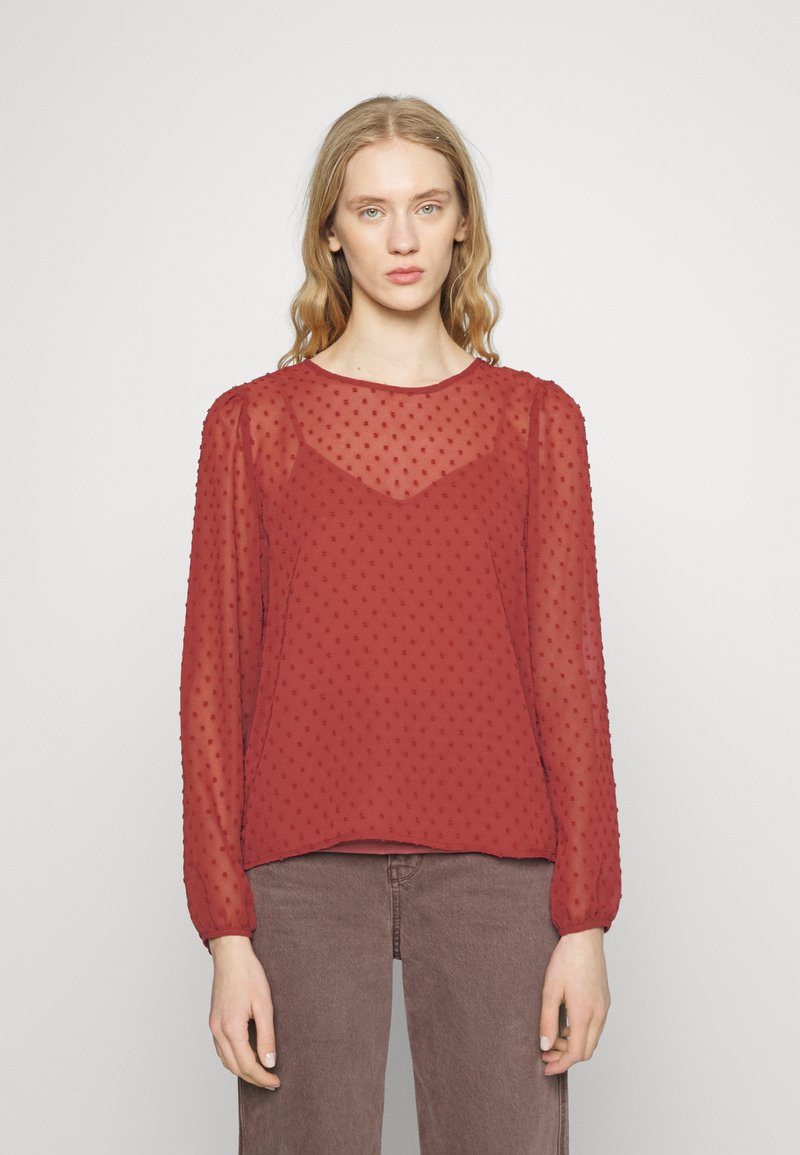 Blusa vermelha transparente com pontos texturizados, gola redonda e mangas longas franzidas nos punhos, combinada com calças de denim castanho.
