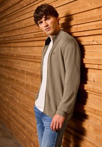 Světle khaki zipová kardigan s žebrovaným límcem, nošený přes bílý tričko. Model stojí proti dřevěné slatě. Viditelné modré džíny.
