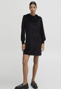 Schwarzes Hoodie-Kleid aus weichem, plüschigem Stoff. Es hat lange Puffärmel, eine Kordelzugkapuze und einen lockeren Schnitt. Kombiniert mit schwarzen Slippern.