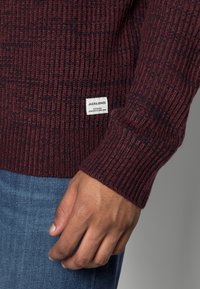 Bordeauxröd ribbad stickad tröja med en lapp som har "JACK & JONES." Texturen är mjuk, och den har långa ärmar, parat med blå denimjeans.