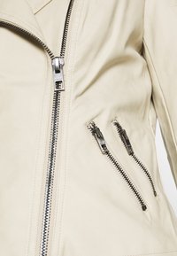 Zbliżenie beżowej skórzanej kurtki z trzema srebrnymi zamkami błyskawicznymi: jednym pionowym z przodu i dwoma ukośnymi zamkami kieszeni na wysokości klatki piersiowej.