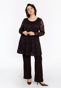 Vrouw staand in een donkerpaarse kanten tuniek met lange mouwen, donkere wijde broek en bruine spitse hakken op een witte achtergrond.