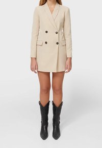 Robe blazer beige croisée avec manches longues, poches avant et boutons noirs, associée à des bottes de cow-boy noires montant jusqu'aux genoux.