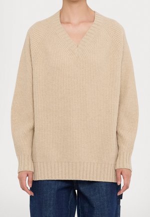 Beiger Strickpullover mit V-Ausschnitt, gerippter Struktur und lockerer Passform. Mit Raglanärmeln und geradem Saum, kombiniert mit dunklen Jeans.