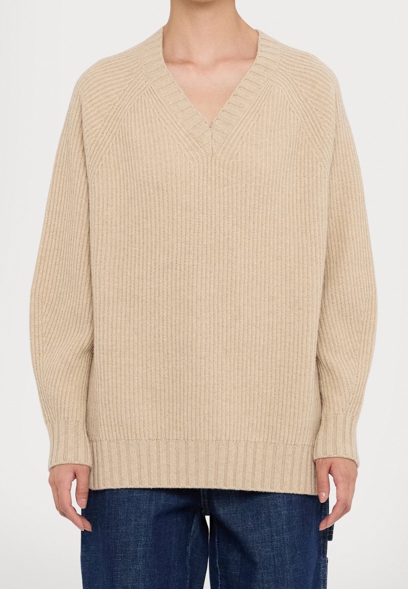 Suéter de punto beige con escote en V, textura acanalada y corte holgado. Presenta mangas ranglán y un dobladillo recto, combinado con unos jeans de mezclilla oscuros.