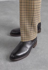 Mustad nahast kingad sileda viimistlusega, ümarate varvastega ja pruuni kihtedega kanna ning houndstooth mustriga pükstega.