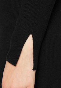 Tissu noir côtelé avec un détail découpé au niveau du poignet, présentant une texture lisse et un ajustement près du corps. La manche dépasse légèrement le poignet.