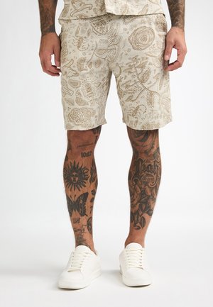 Beżowe szorty z wzorem kwiatowym i paisley, wykonane z lekkiego materiału. Połączone z białymi tenisówkami.
