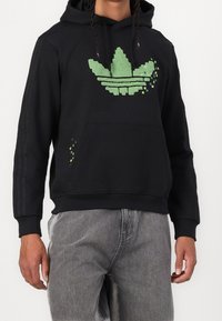 Svart hoodie med grön pixellogga, känguruficka, ribbade muddar och tre svarta ränder längs ärmarna.