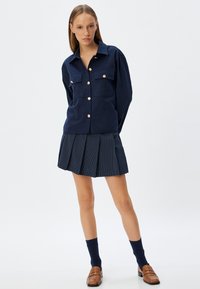 Camicia a maniche lunghe blu navy con due tasche sul petto e bottoni dorati, abbinata a una gonna plissettata con motivi a righe. Mocassini marroni e calze.
