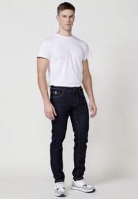 Jeans de mezclilla de un azul oscuro con costuras amarillas, combinados con una camiseta blanca ajustada. El modelo lleva zapatillas blancas y grises.