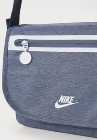 Borsa messenger in tessuto blu con tasca con zip bianca e logo Nike bianco. Caratterizzata da una finitura testurizzata e una tracolla regolabile.