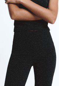 Keresztbe font karral álló személy fekete, csillogó, testhez simuló nadrágot és hozzáillő felsőt visel, egyszínű, világos háttér előtt.