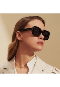 Schwarze übergroße Sonnenbrille mit dicken Rahmen, die ein glattes Finish und ein goldenes Logo an den Bügeln aufweist. Tragen Sie sie mit einer hellen Jacke.