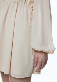 Helles beigefarbenes Kleid mit geraffter Taille, weichem Stoff und langen, weit schwingenden Ärmeln. Verfügt über eine Kordelzugdetails und einen leicht ausgestellten Saum.