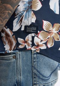 Camicia a maniche corte a fiori con base blu scuro, grandi fiori color crema e marrone, e etichetta "Petrol Ind". Abbinata a jeans di denim chiaro.