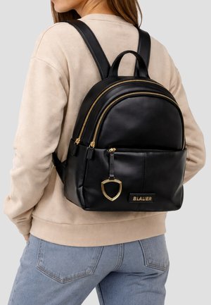 Donna che indossa una felpa beige e jeans blu con uno zaino in pelle nero con cerniere dorate e il logo Blauer sul retro.