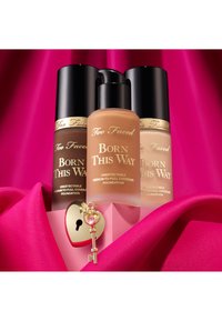 Drie cilindervormige flessen van de foundation "Born This Way" in verschillende tinten, met een gouden sleutel en een hartvormig slot op roze stof.