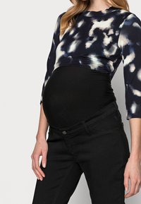 Persona embarazada vistiendo una blusa de maternidad negra y pantalones negros con una camisa de manga larga azul y blanca con un patrón.