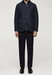Man in een donkerblauwe blazer over een grijze ziptrui, donkere broek en zwarte dresschoenen, staande tegen een effen achtergrond.
