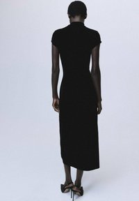 Vestido de terciopelo negro con mangas cortas, silueta entallada, longitud midi y cuello alto. Llevado con sandalias negras de tacón alto.