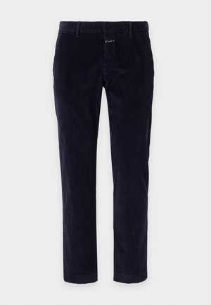 Hombre que lleva una camiseta polo blanca de manga corta, pantalones oscuros y mocasines de cuero marrón, de pie con una mano en el bolsillo sobre un fondo claro.