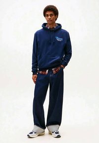 Marineblauw hooded sweatshirt met logo, gedragen over een rood-wit geruit overhemd. Losvallende spijkerbroek en sneakers met patroon.