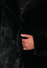 Hånd med manikurerede negle holder den lukkede krave af en blød sort faux fur frakke, der afslører en sort blondetop nedenunder.