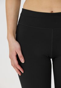 Svarta stretch leggings med hög midja, som har en slät yta och subtila sömdetaljer, visade med en hand som vilar på låret.