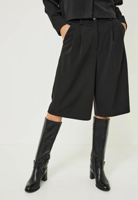 Černé přiléhavé šortky vyrobené z hladkého materiálu s záhyby, doplněné černými koženými holinami pojímanými na vysokém podpatku a s akcentovým páskem.