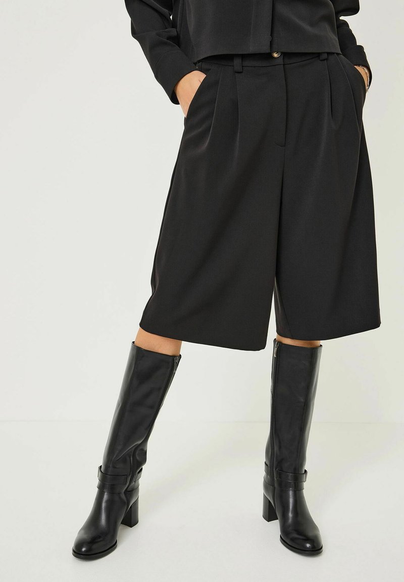 Černé přiléhavé šortky vyrobené z hladkého materiálu s záhyby, doplněné černými koženými holinami pojímanými na vysokém podpatku a s akcentovým páskem.