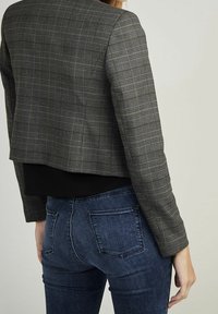 Blazer cropped a quadri grigi con design aderente, abbinato a una maglia nera e jeans skinny blu scuro. Vista dal retro che mostra i dettagli.
