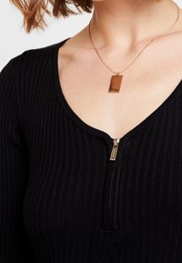 Zwarte geribbelde trui met een rits bij de hals, voorzien van een gouden rechthoekige hangersketting. Zacht, gestructureerd materiaal.