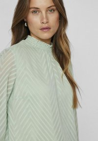Light green blouse with a textured chevron pattern, a smocked neckline, and long sleeves. Material appears soft and lightweight.