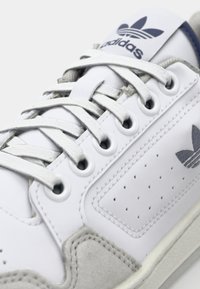 Zapatilla blanca con detalles perforados y acentos de ante gris. Presenta cordones blancos, un logo en la lengüeta y un acabado de cuero suave.