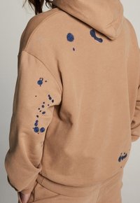 Sweat à capuche en tissu beige avec broderies bleues et motifs circulaires au dos et sur la manche droite, présentant une coupe décontractée.