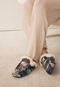 Pantofole con motivo floreale su base blu navy e fodera in pelliccia sintetica; design a punta; tessuto visibile; indossate con pantaloni beige chiaro.