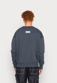 Mörkgrå tröja med avslappnad passform, ribbade ärmmuddar och nederkant. Anmärkningsvärd detalj: rektangulär patch på övre delen av ryggen. Mjuk tygtextur.