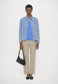 Denim jacket with cropped hem, light blue; blue sweater underneath; beige pants; black handbag; black flat shoes; model standing upright.