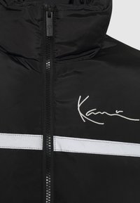 Svart vadderad jacka med hög krage, dragkedja framtill och vit horisontell rand. Inkluderar broderad logotyp i silvertråd.