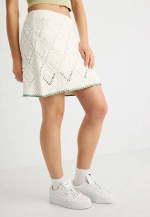 Witte gebreide rok met een diamantpatroon en ajourdetails, afgewerkt met een groene accent aan de zoom. Draagt witte sneakers.